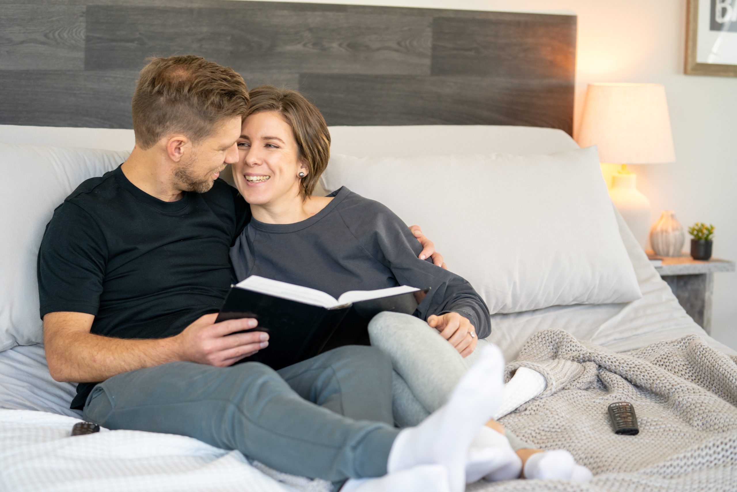 A couple smiles while reading a book on a split adjustable bed, which can help find a comfortable position for how to sleep in the heat.