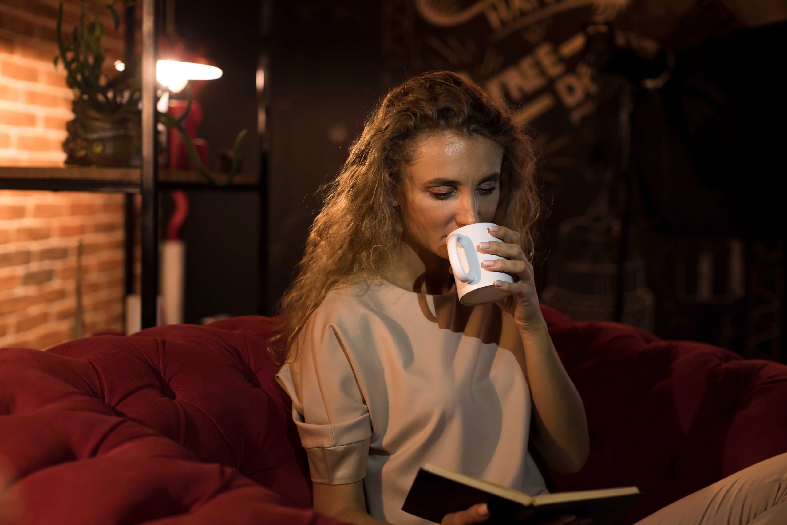 A woman practicing a calming wind-down routine by reading and drinking a warm beverage in dim lighting to signal safety to her nervous system.
