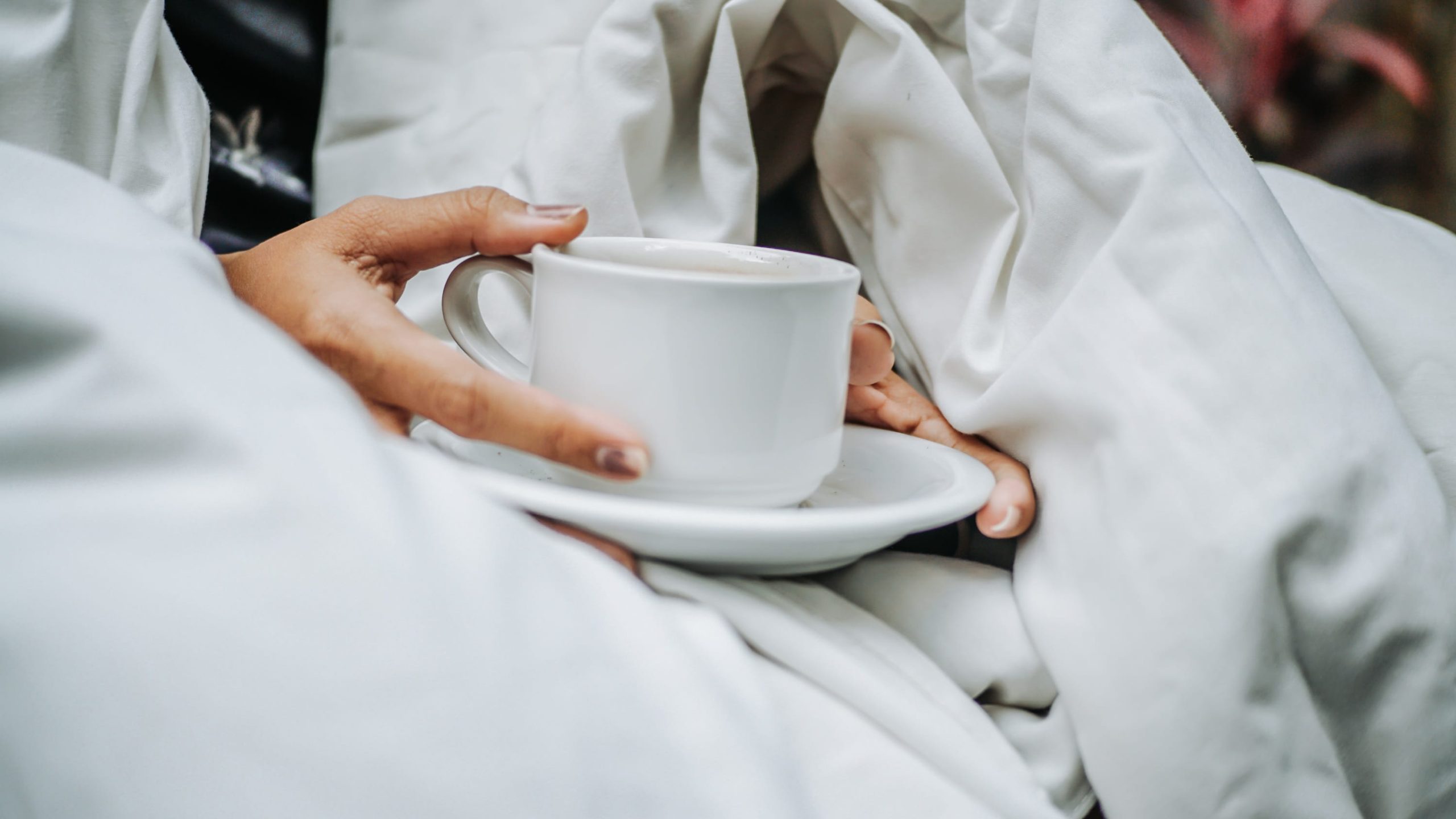 Hands holding a cup of coffee in bed — a common trigger for nighttime acid reflux