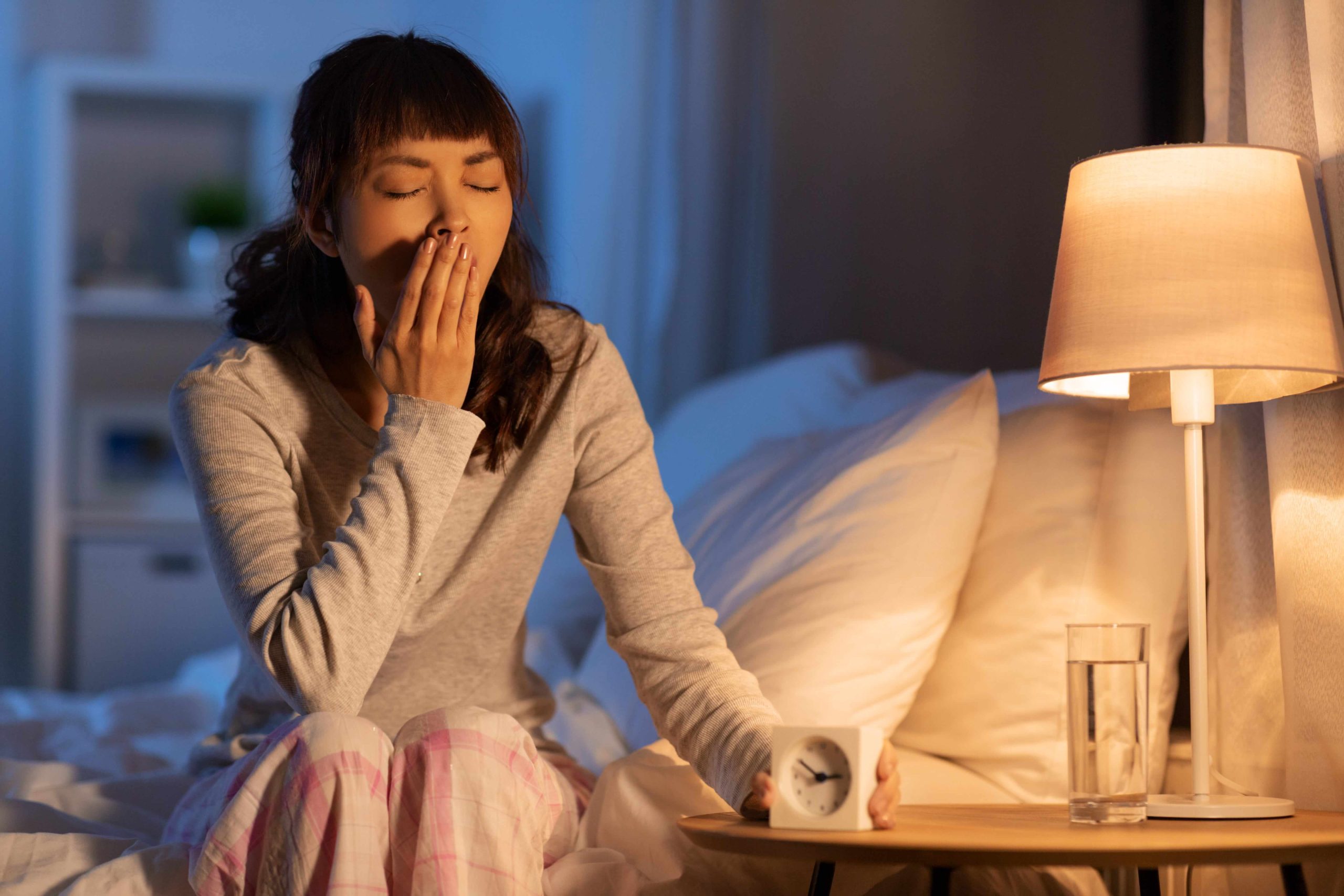 An exhausted woman yawning in bed next to a lamp and alarm clock, highlighting the fatigue caused by disrupted sleep.
