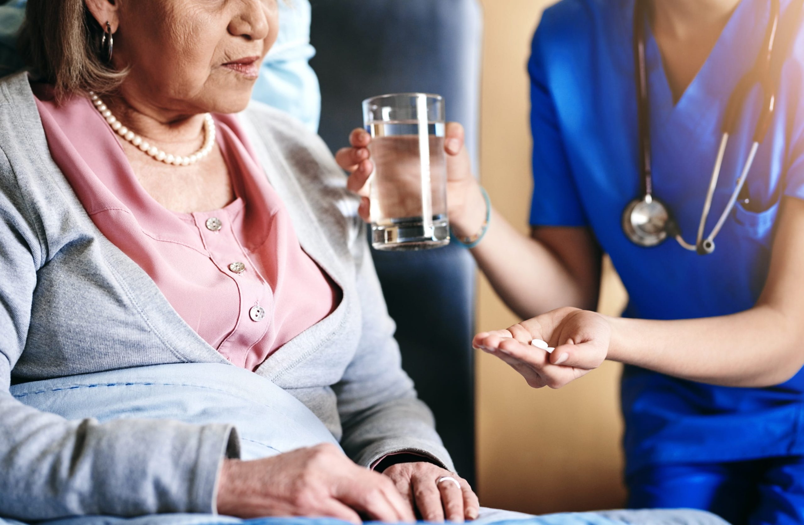 A close-up of a healthcare professional in blue scrubs handing a glass of water and medication to an older patient