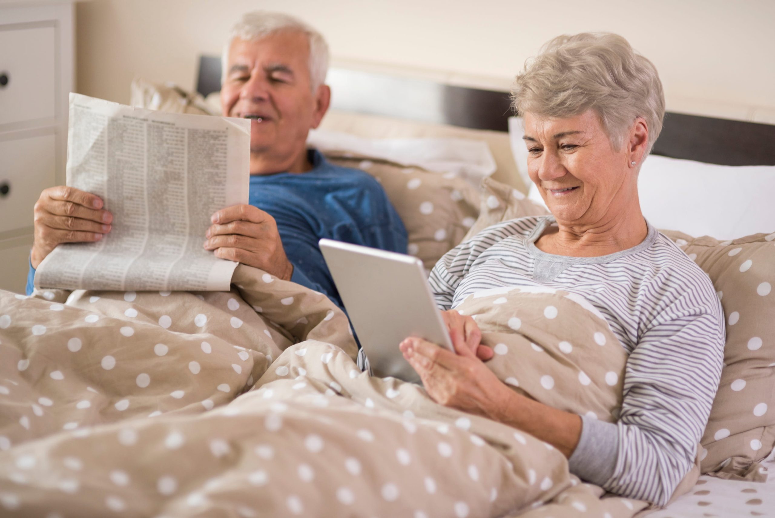 An older man and woman sitting up in bed during a relaxed morning or evening; the man is reading a newspaper and the woman is using a digital tablet.